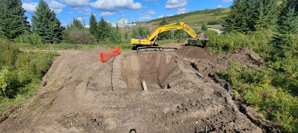 An excavator is situated atop a mound of dirt, with an orange safety barrier present nearby. The excavator's yellow colora...